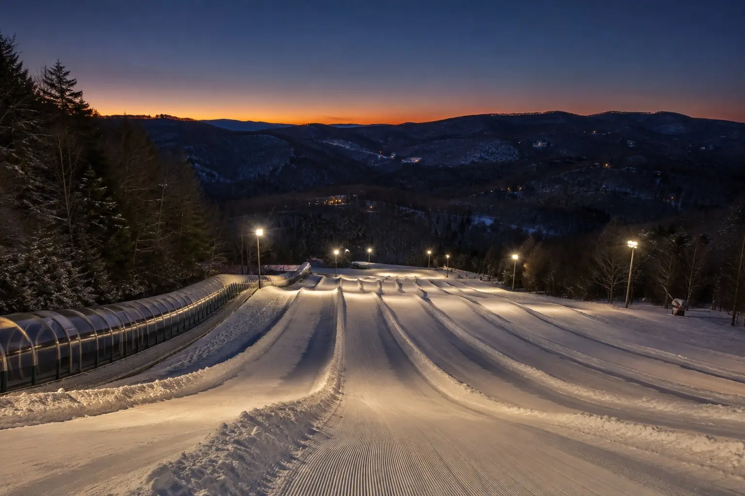 Floodlit tubing lanes of groomed snow running downhill at dusk, with a covered conveyor lift alongside them and layered mountain ridges under an orange band of sunset in the distance.