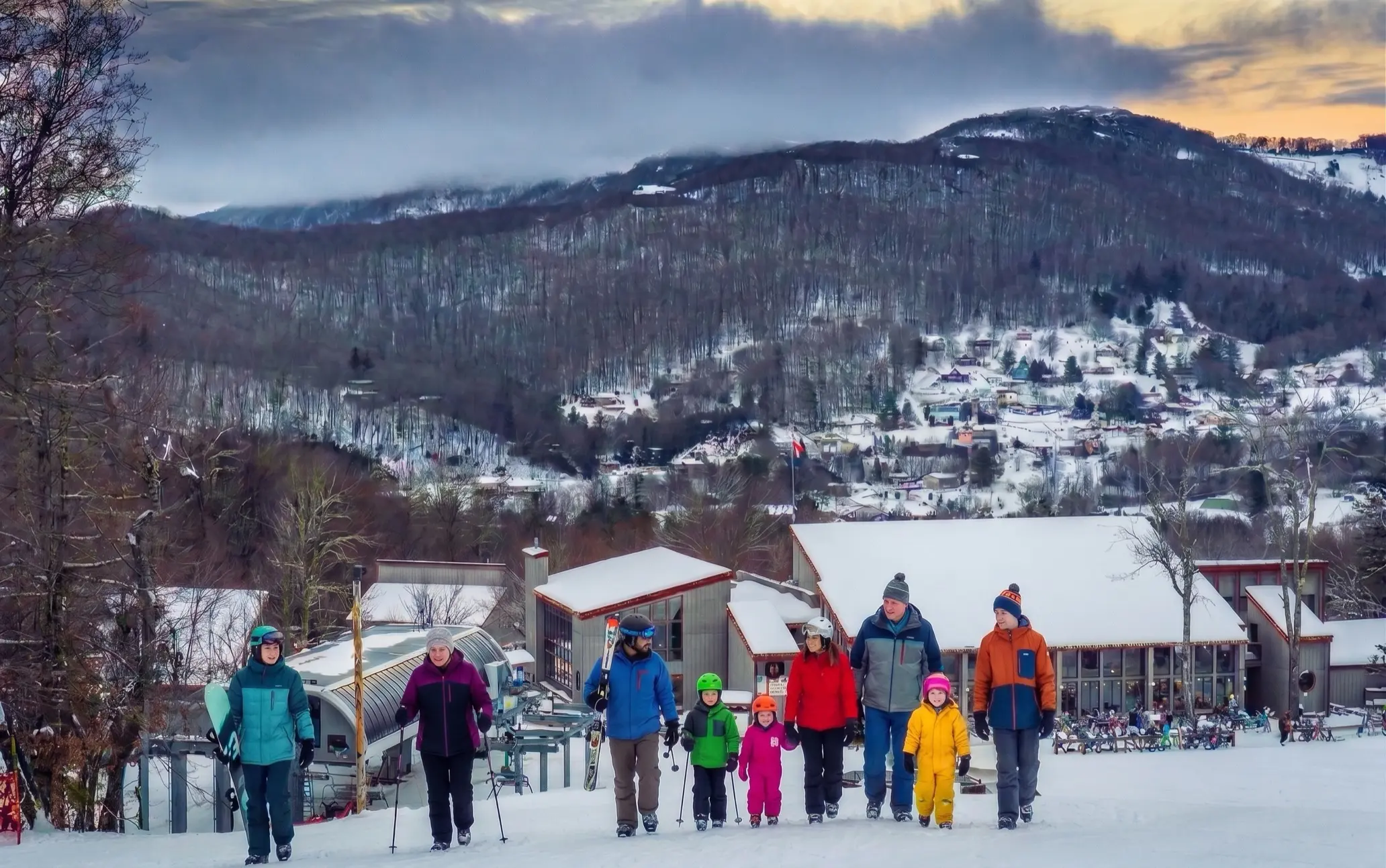 Skiers and families of mixed ages walking down a snowy slope toward the base area buildings at dusk, with forested ridges and scattered houses on the hillside behind