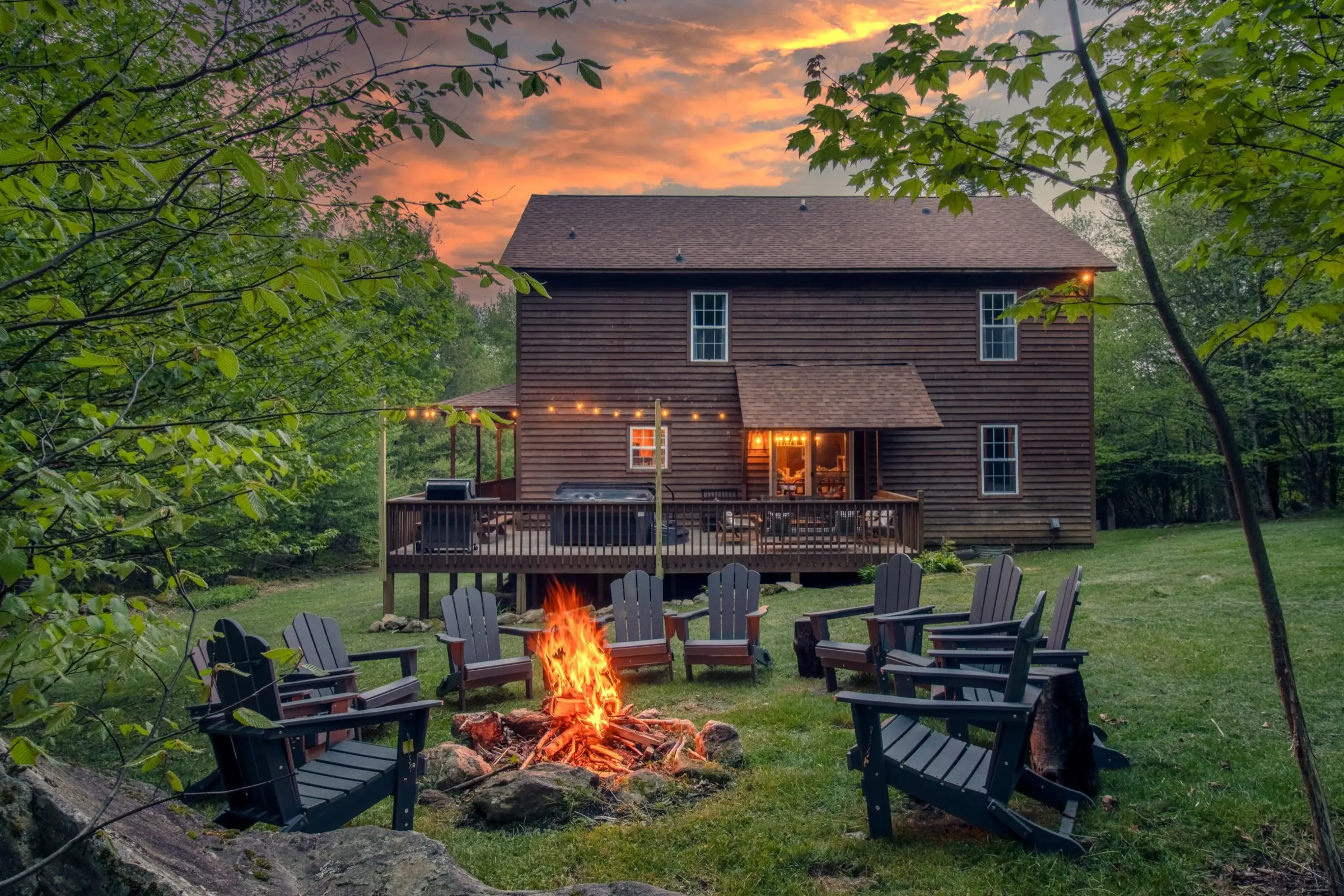 The Sugar Nest at sunset. A fire pit burns in the foreground ringed by Adirondack chairs, with the lit rear deck, hot tub and string lights behind.